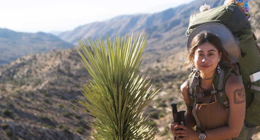 A person wearing a backpack pauses for the photo beside a desert plant. In the background, there are mountains. 